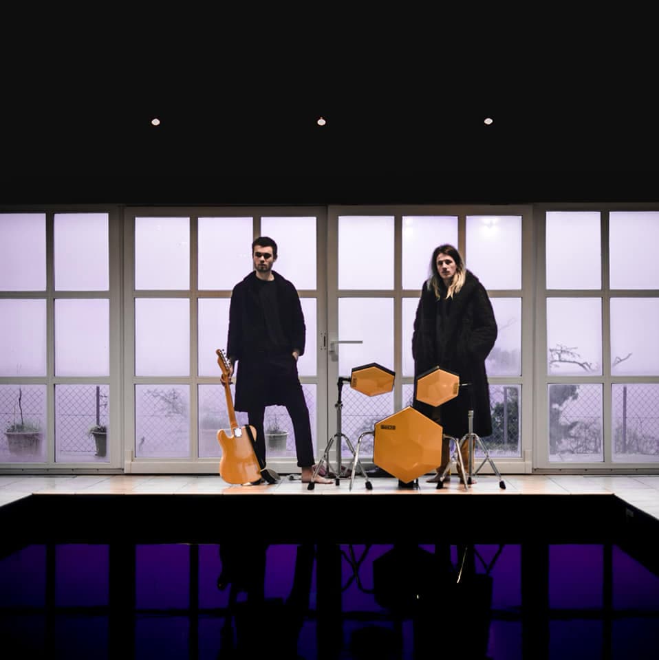 Deux musiciens de Chemical Fame sur scène, l'un avec une guitare orange, l'autre devant des tambours jaunes géométriques, devant une baie vitrée
