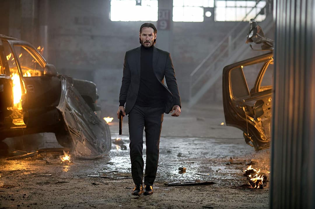 John Wick en costume noir et arme à la main marche dans un entrepôt avec des voitures en feu