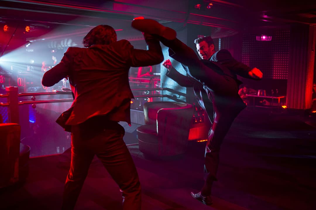 Keanu Reeves dans une scène d'action du film John Wick, effectuant un coup de pied acrobatique dans un décor futuriste aux néons rouges et roses