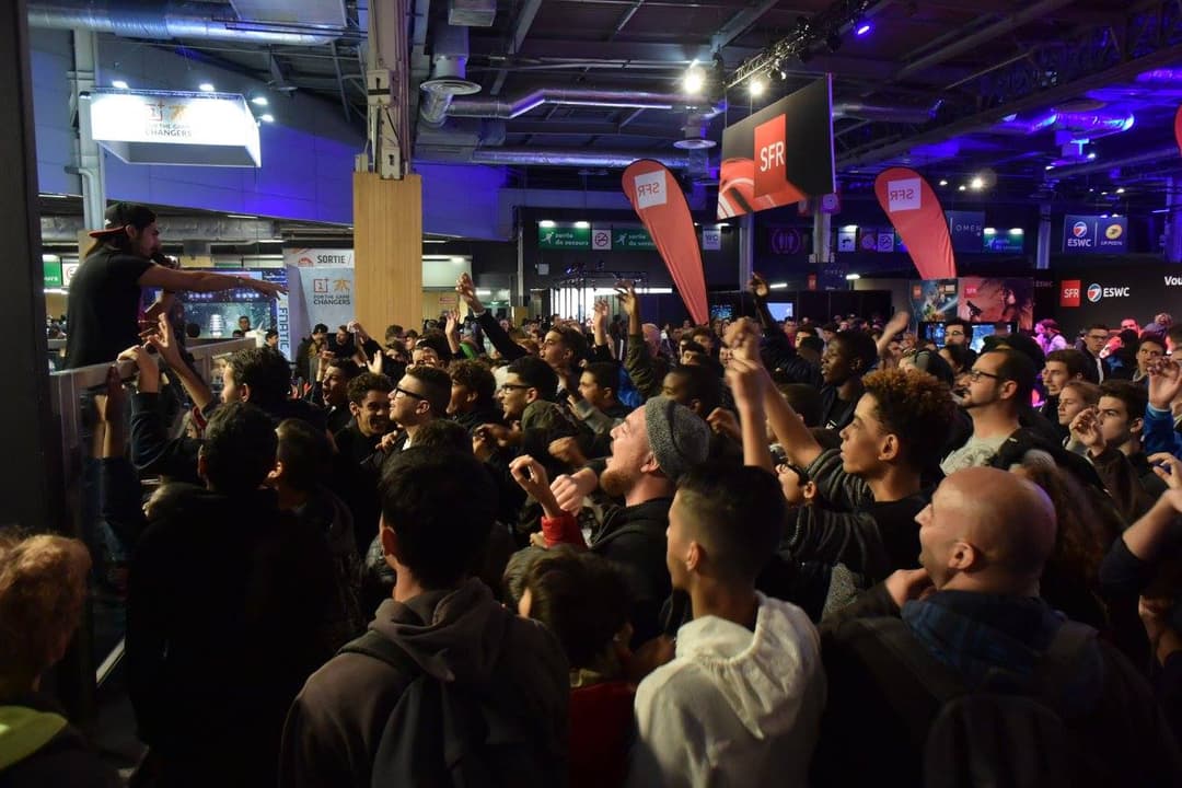 Foule de visiteurs dense à la Paris Games Week, éclairage bleu et orange, stands et écrans de jeux vidéo en arrière-plan