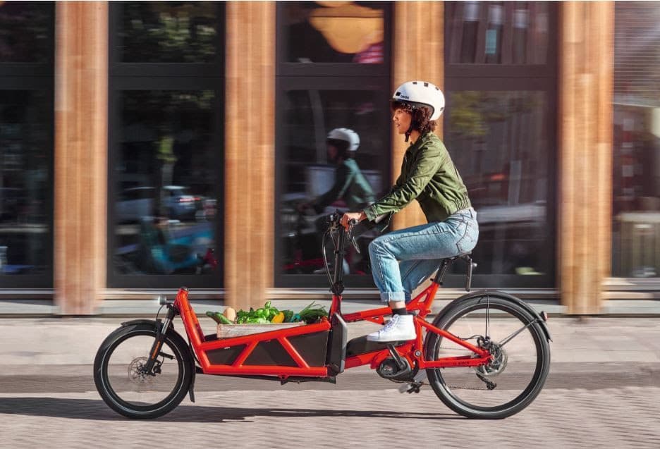 Femme à vélo électrique rouge cargo en milieu urbain, portant un casque blanc et une veste verte