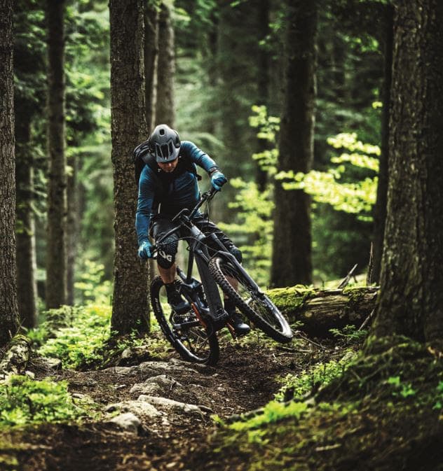 Cycliste en vélo électrique VTT roulant sur un sentier forestier entre les arbres, vêtu d'un gilet bleu et d'un casque blanc