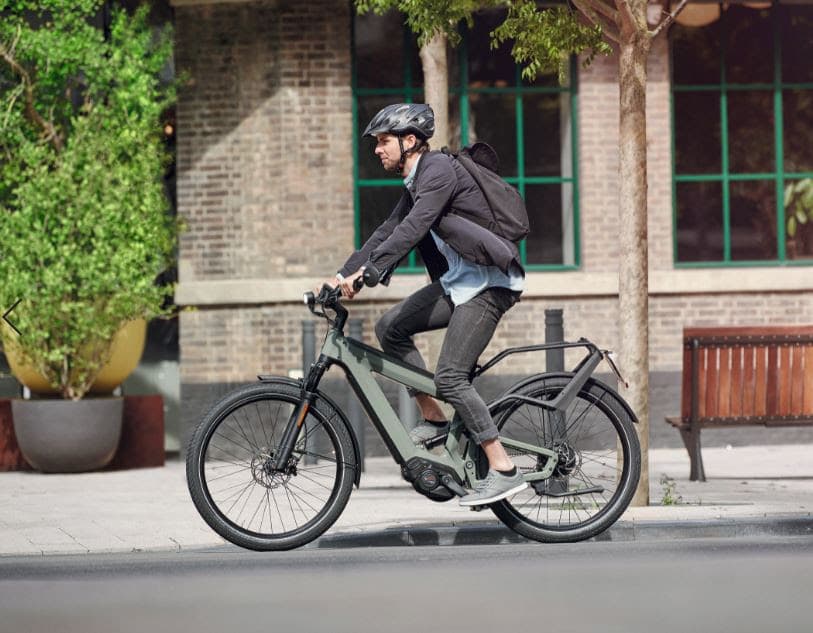 Cycliste portant un casque et une veste noire chevauchant un vélo électrique gris en milieu urbain