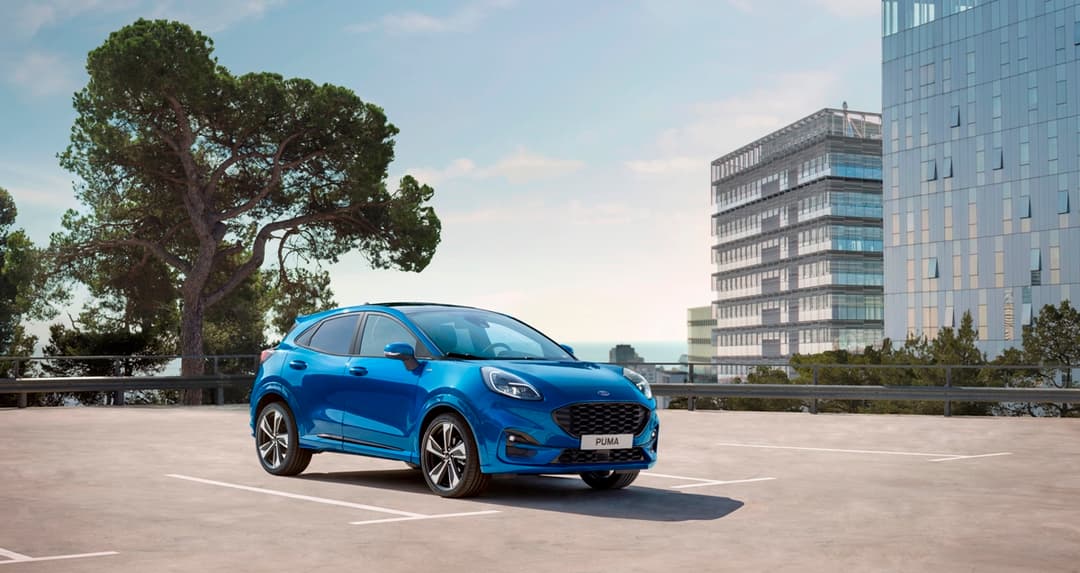 Ford Puma bleu garé sur un parking, avec immeuble moderne et grand arbre en arrière-plan