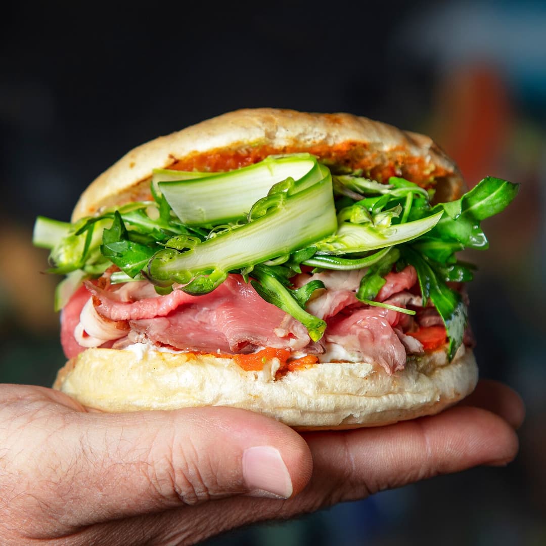 Burger sur focaccia garni de burrata, tomates concassées, roquette et asperges vertes, tenu dans une main