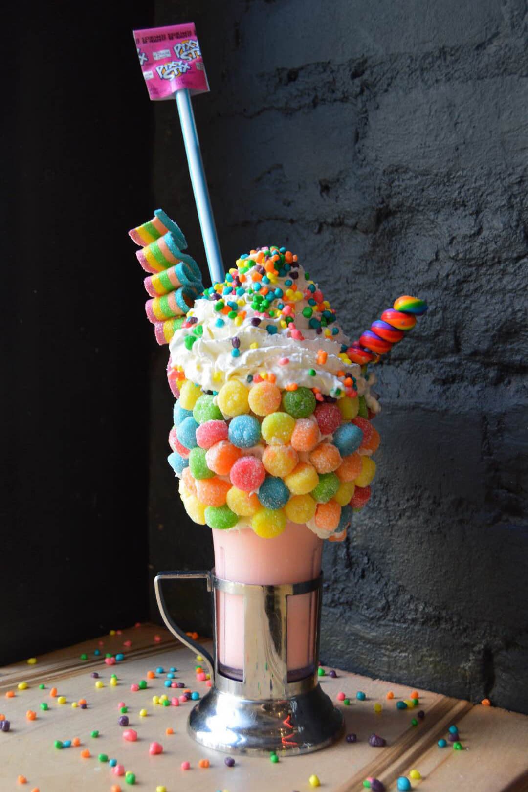 Crazy shake Black Tap coloré recouvert de bonbons multicolores, serpentins de bonbons et drapeau rose, sur fond de mur gris
