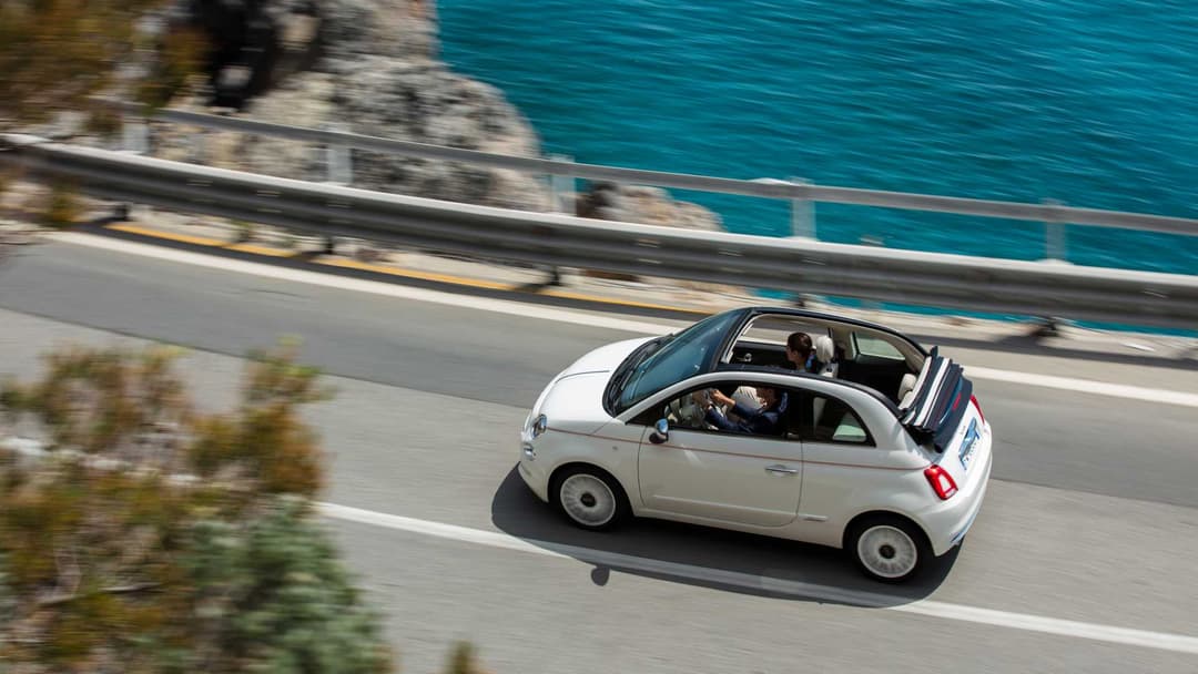 Fiat 500 Dolcevita blanche décapotée roulant sur une route côtière avec mer turquoise en arrière-plan