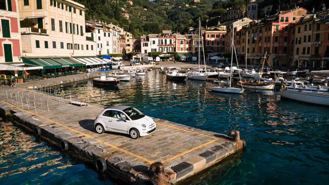 Fiat 500 Dolcevita blanche garée sur un quai face à un port méditerranéen avec voiliers et façades colorées