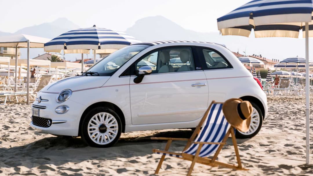 Fiat 500 Dolcevita blanche garée sur une plage avec parasols bleu et blanc et transat en premier plan