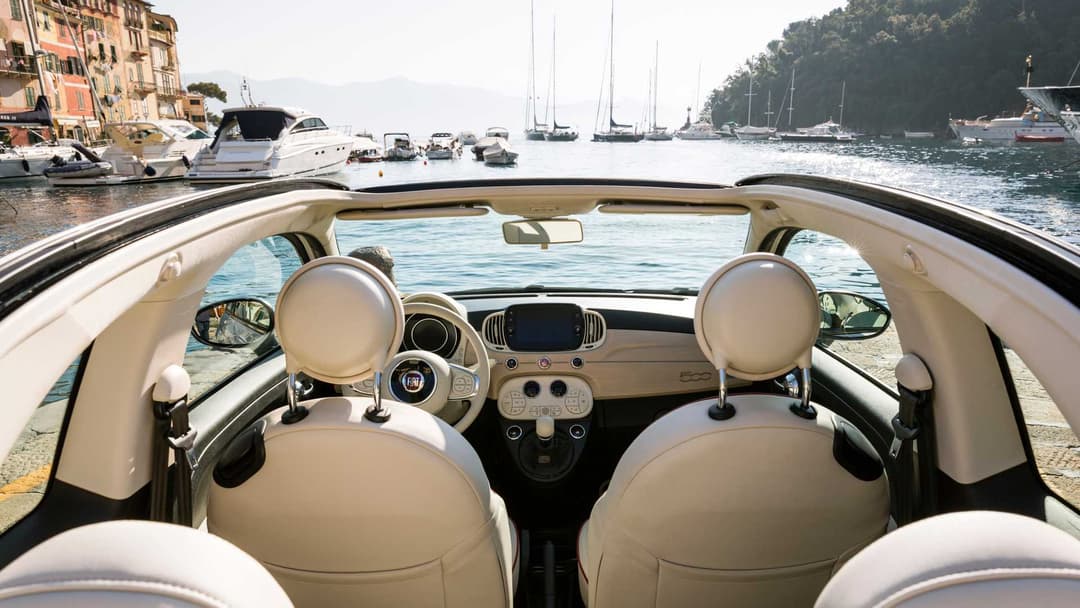 Fiat 500 Dolcevita décapotable blanc crème, vue de l'intérieur avec volant et sièges beige, face à une baie méditerranéenne avec bateaux
