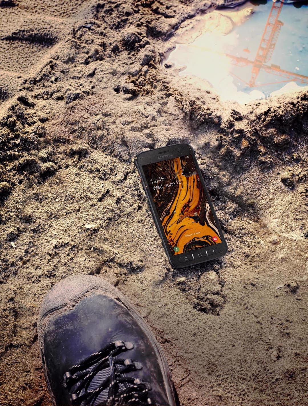 Samsung Galaxy XCover 4s posé sur le sable et les cailloux d'un chantier, avec une botte de travail au premier plan