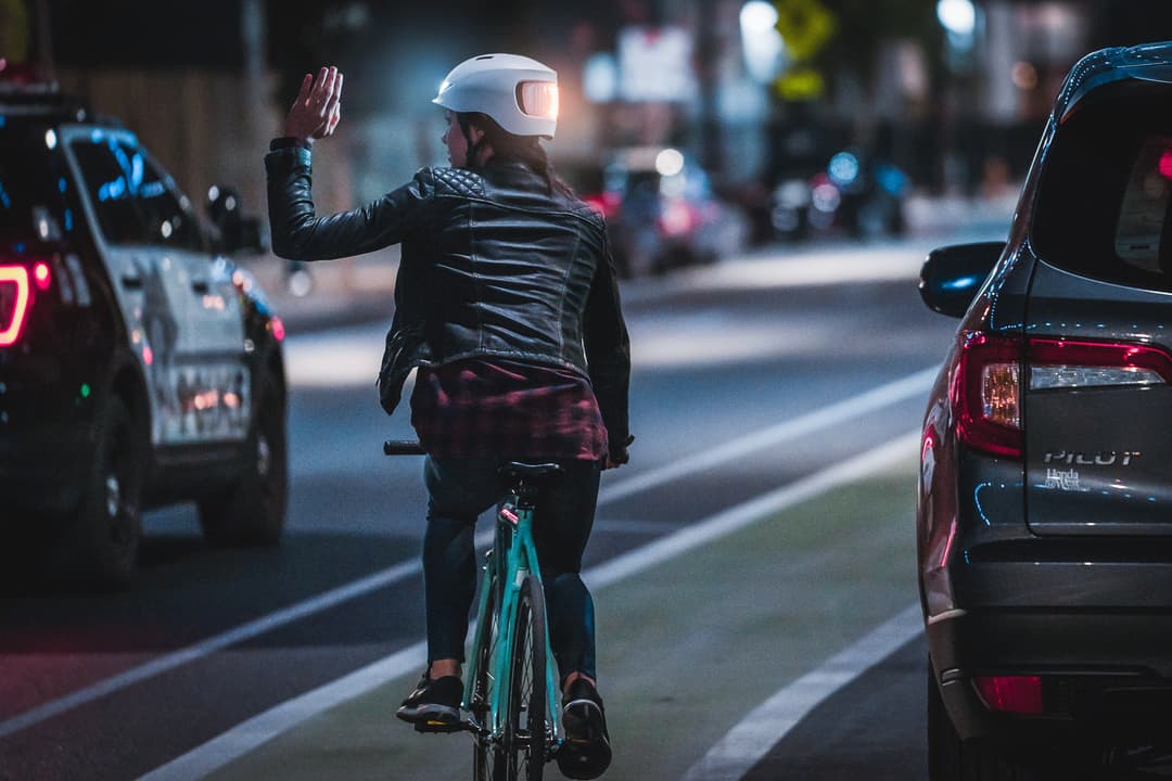 Cycliste de nuit portant un casque Lumos blanc avec LED illuminées, levant la main dans un carrefour urbain entre voitures