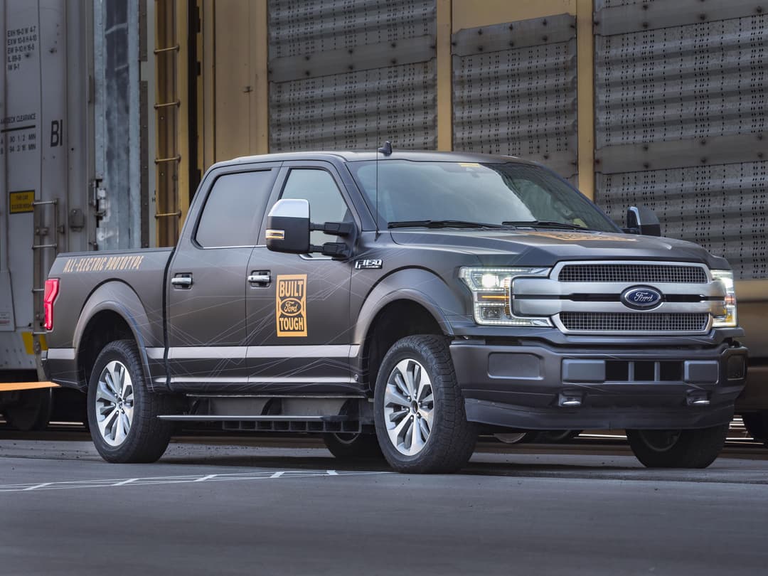 Ford F-150 électrique gris foncé de profil, avec badge « Built Ford Tough », stationné face à des wagons de transport