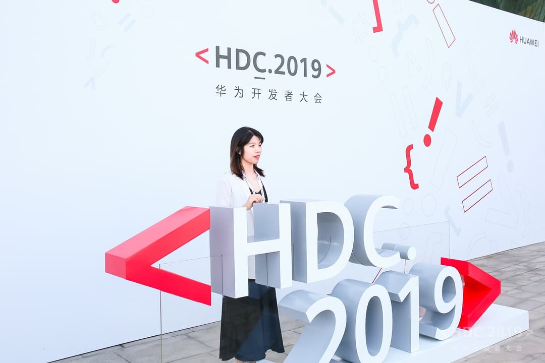 Femme devant la signalisation de la Huawei Developer Conference 2019, avec logo HDC.2019 en blanc et rouge sur fond blanc