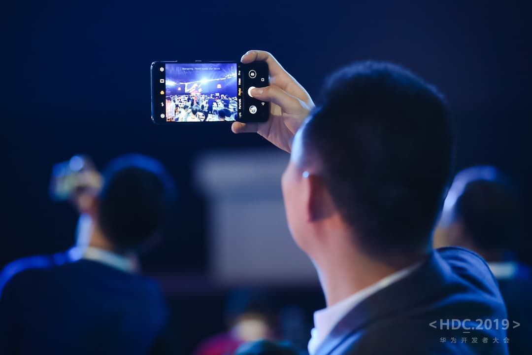 Main tenant un smartphone Huawei en mode horizontal pour photographier une scène de conférence éclairée en bleu, lors de la HDC 2019