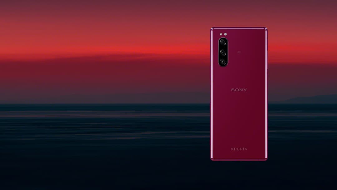 Sony Xperia 5 de couleur rouge bordeaux, vue de face, présenté devant un coucher de soleil sur l'océan