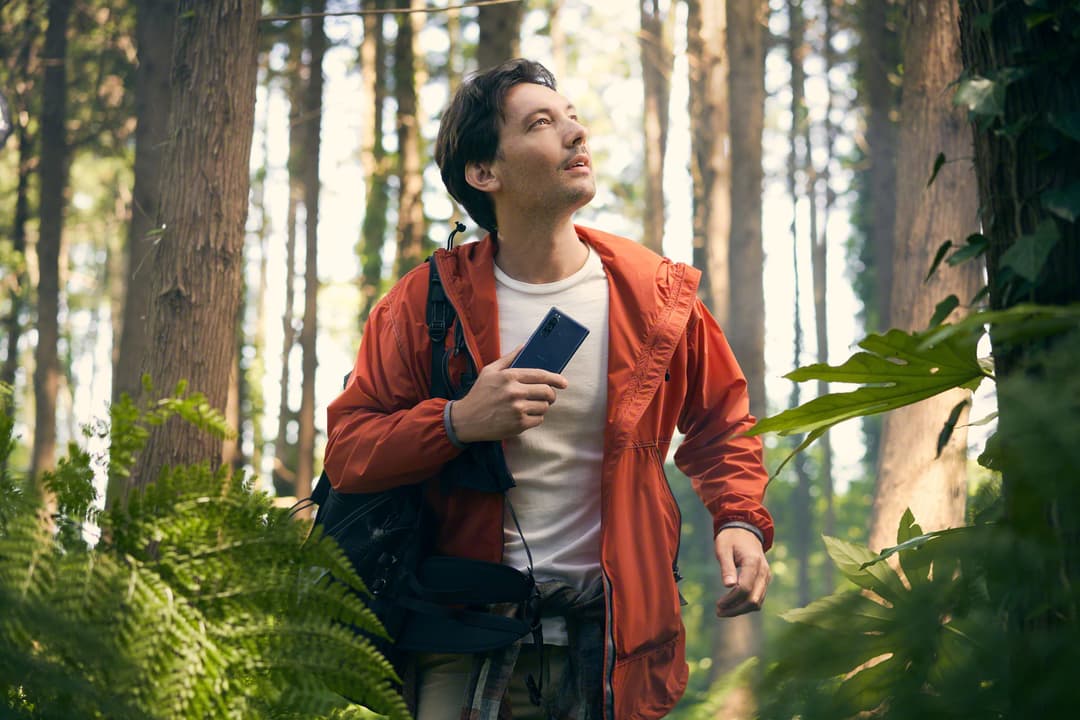 Homme en veste orange tenant un Xperia 5 noir dans une forêt ensoleillée