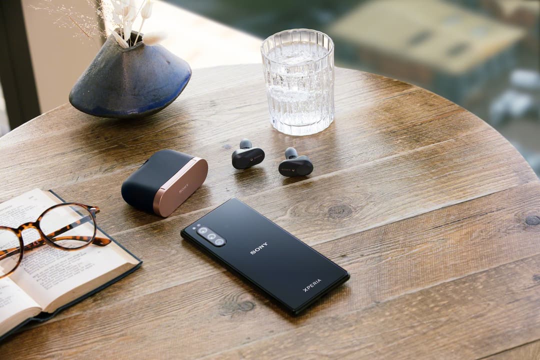Sony Xperia 5 noir posé sur une table en bois avec ses accessoires : écouteurs, boîtier de charge, verre d'eau et lunettes
