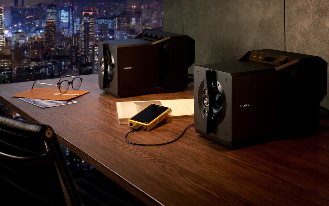 Enceintes Sony SA-Z1 noires posées sur un bureau en bois, avec un téléphone or relié par câble, lunettes et documents à proximité, devant une baie vitrée avec vue sur gratte-ciel illuminés la nuit