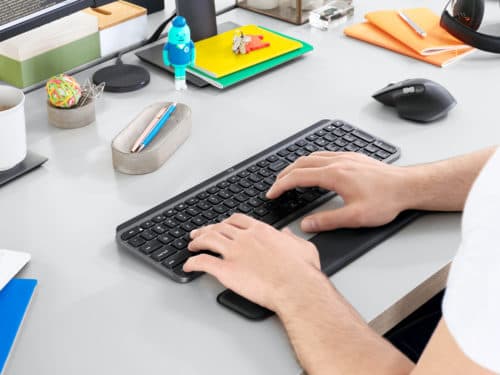 Mains utilisant le clavier MX Keys noir et la souris MX Master 3 noire sur un bureau blanc organisé avec accessoires de travail