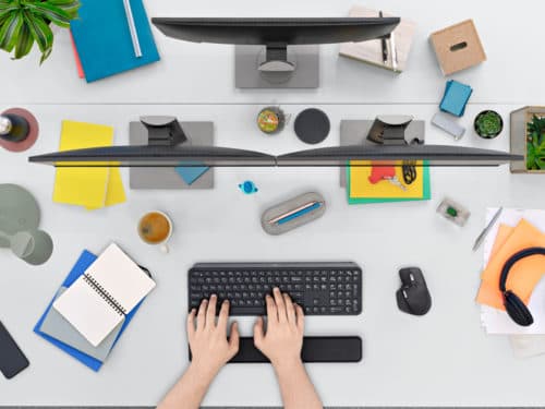 Dessus d'un bureau avec clavier MX Keys noir et souris MX Master 3, mains dactylographiant, écrans multiples et accessoires variés