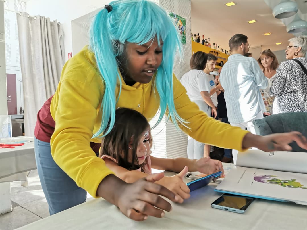 Enfant portant une perruque bleue utilisant une tablette tactile avec l'aide d'une femme en gilet jaune, lors d'une démonstration de technologie