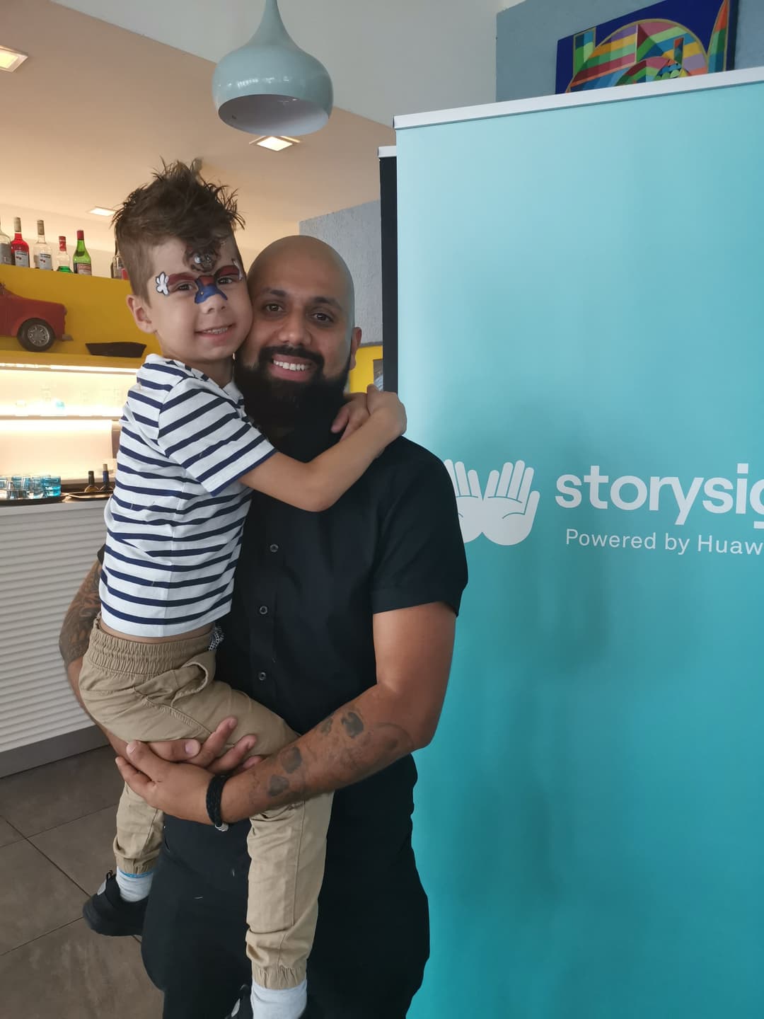 Un homme tenant un enfant dans les bras devant un panneau Huawei StorySign turquoise, dans un intérieur moderne