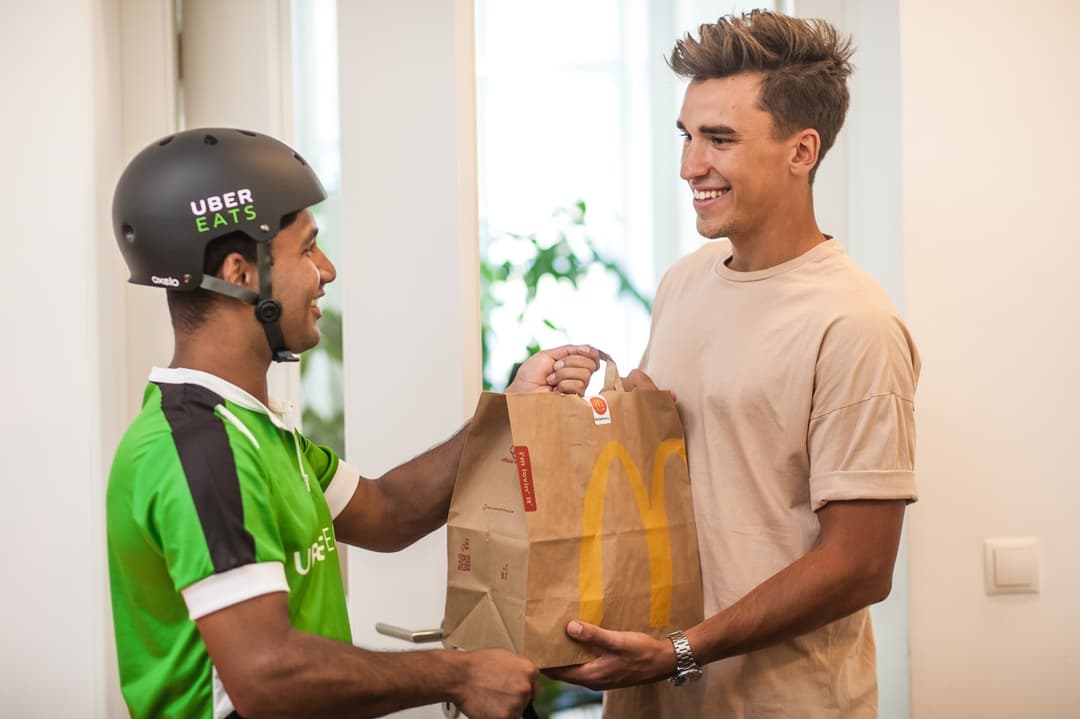 Livreur Uber Eats en gilet vert et casque noir remettant un sac McDonald's à un client souriant à son domicile