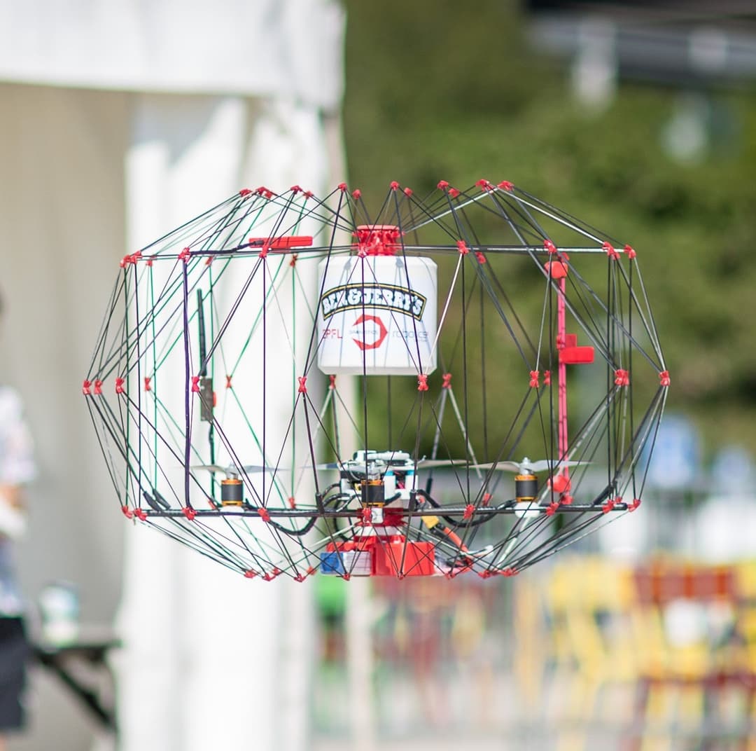 Drone de livraison Ben & Jerry's en vol, avec un pot de glace suspendu sous sa structure de tubes et câbles rouges et gris
