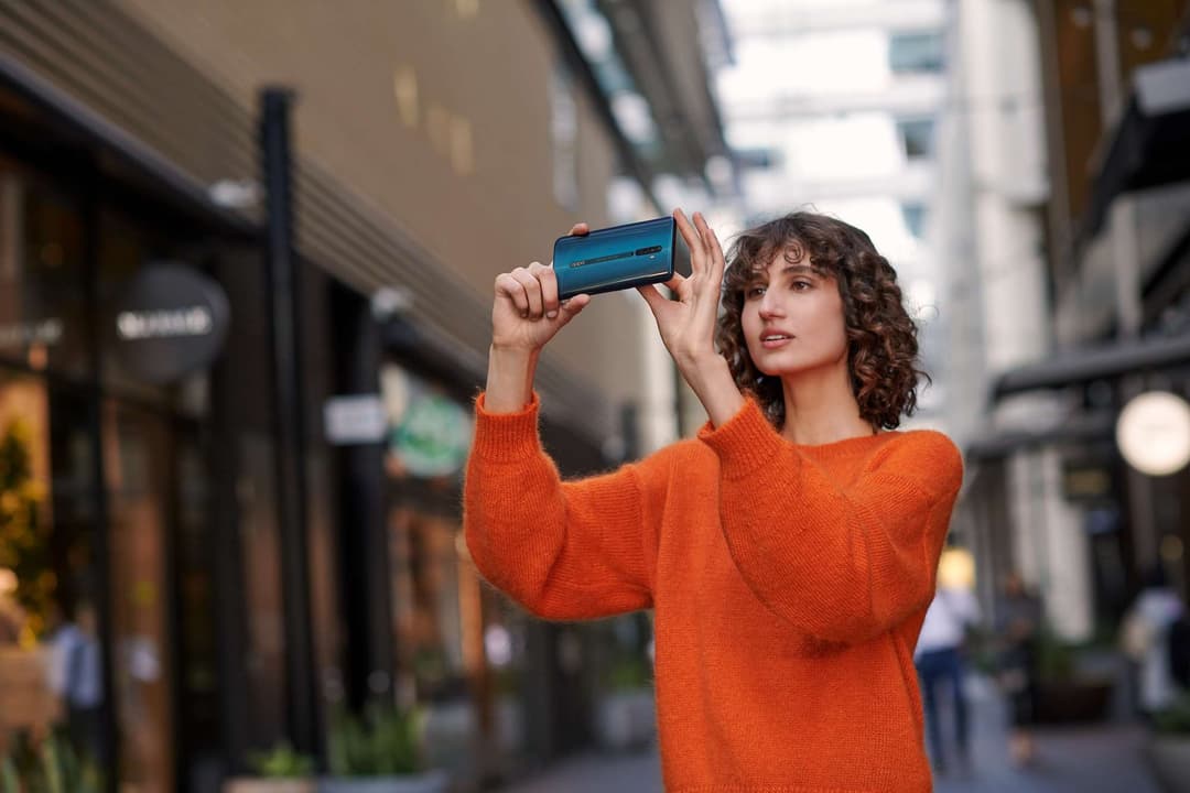 Femme en pull orange tenant un smartphone OPPO Reno2 bleu en mode paysage, dans une rue urbaine