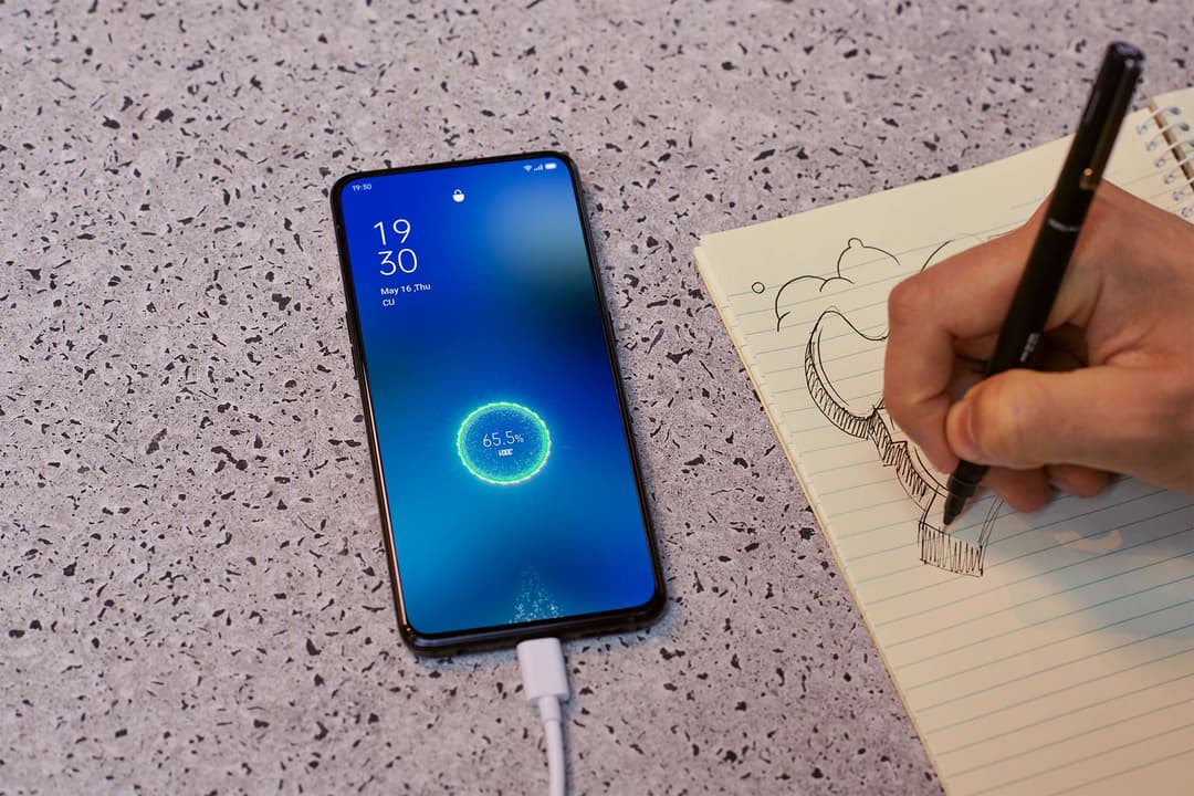 OPPO Reno2 en charge affichant un écran bleu avec indicateur de batterie, aux côtés d'une main dessisant dans un carnet