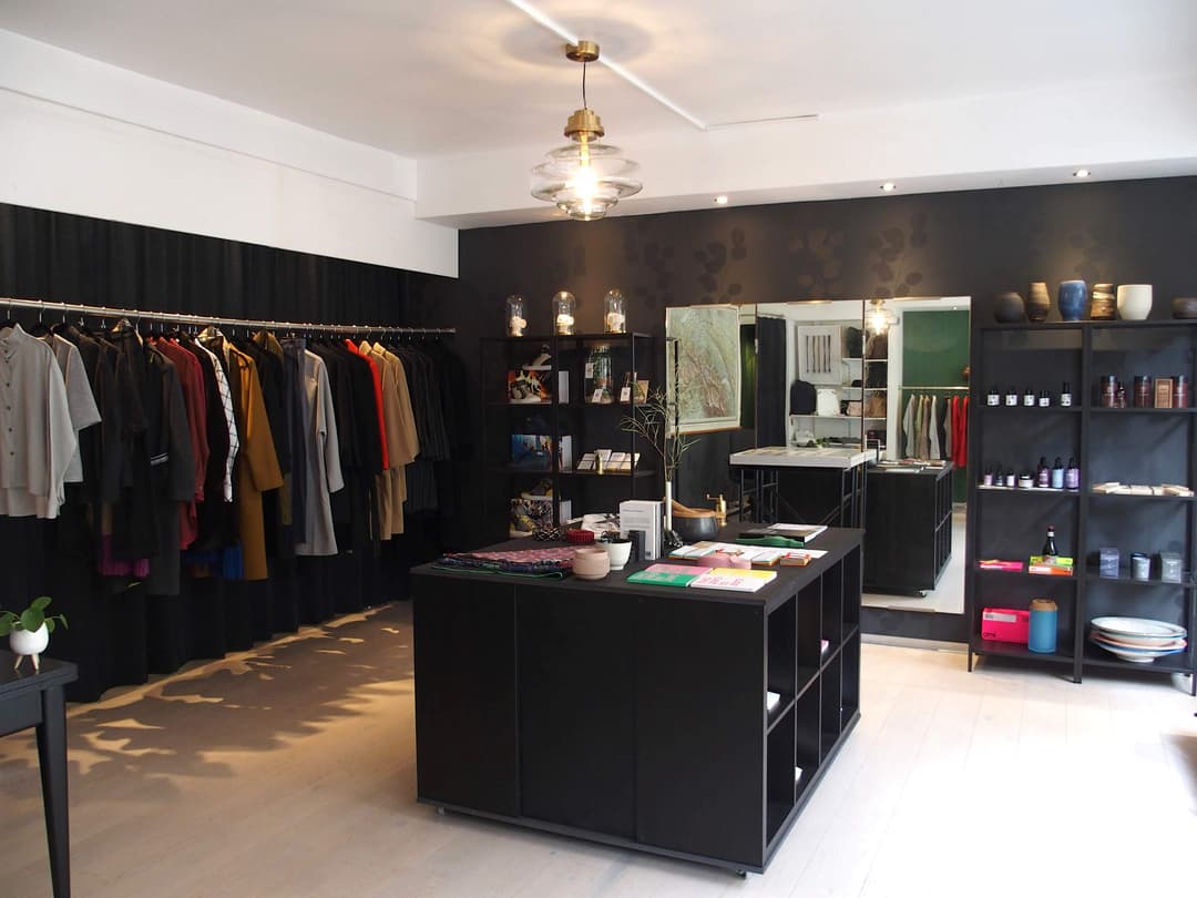 Intérieur du JSBG Store présentant des vêtements sur portant, mobilier noir épuré, étagères de produits artisanaux et luminaires design