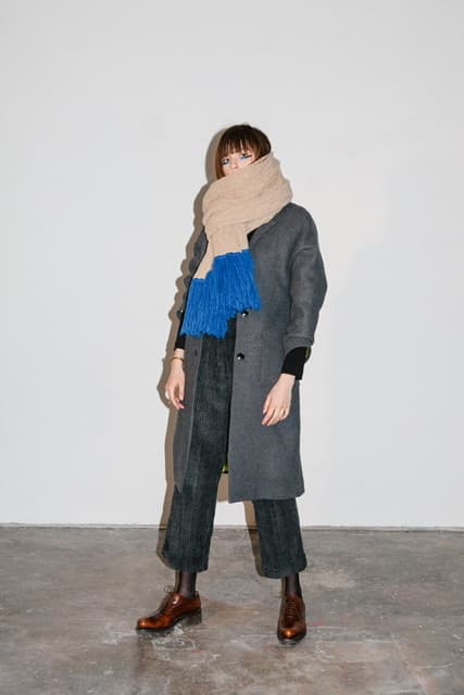 Femme portant un manteau gris, un foulard beige, un pull bleu et un pantalon noir, création suisse indépendante présentée au JSBG Store