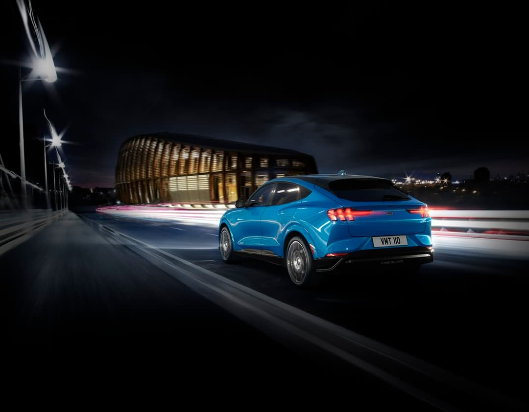 Ford Mustang Mach-E bleu électrique de profil, circulant de nuit devant un bâtiment moderne illuminé