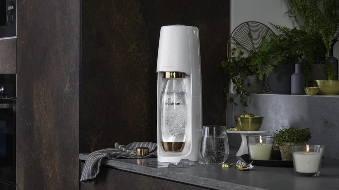 Appareil Sodastream Gold Edition blanc et or sur un plan de travail de cuisine, entouré de bougies et plantes décoratives