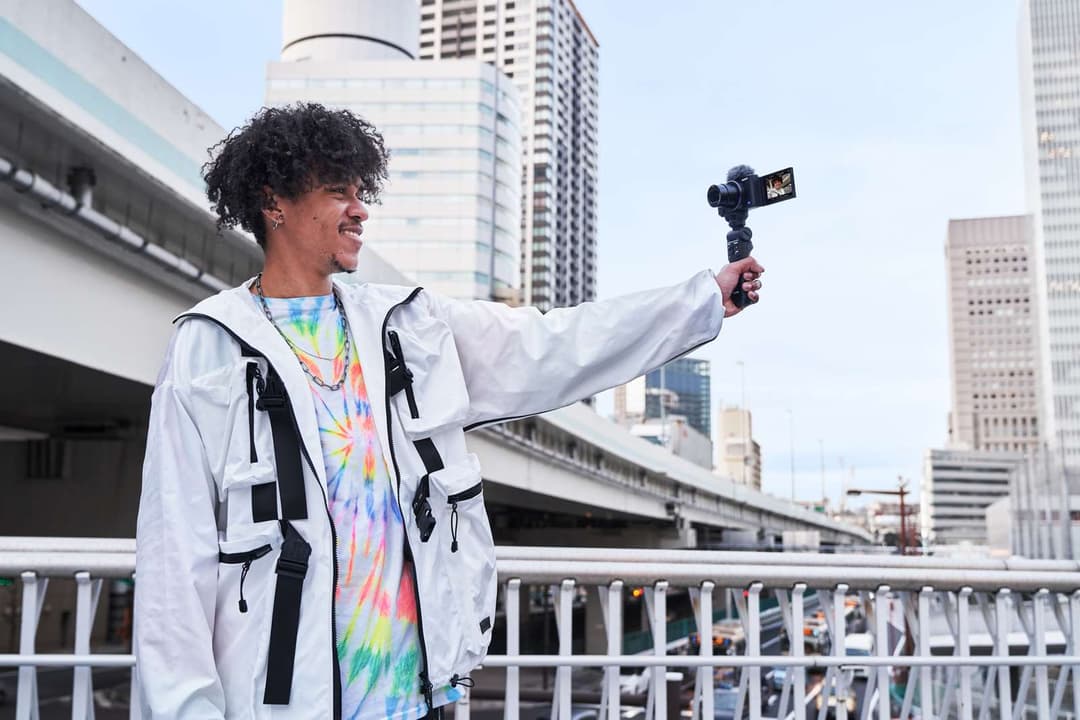Jeune homme filmant avec l'appareil photo compact Sony ZV-1 en selfie, écran LCD orienté vers l'avant, en veste blanche sur un balcon urbain