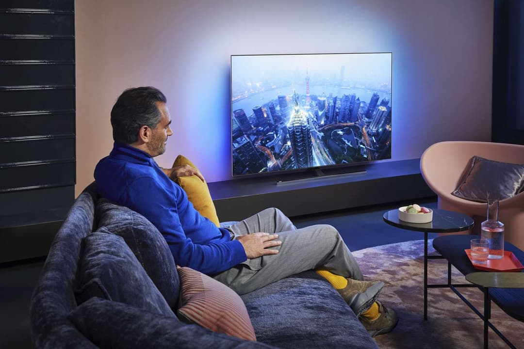 Homme assis sur un canapé regardant un téléviseur Philips OLED affichant une vue aérienne de gratte-ciels illuminés la nuit