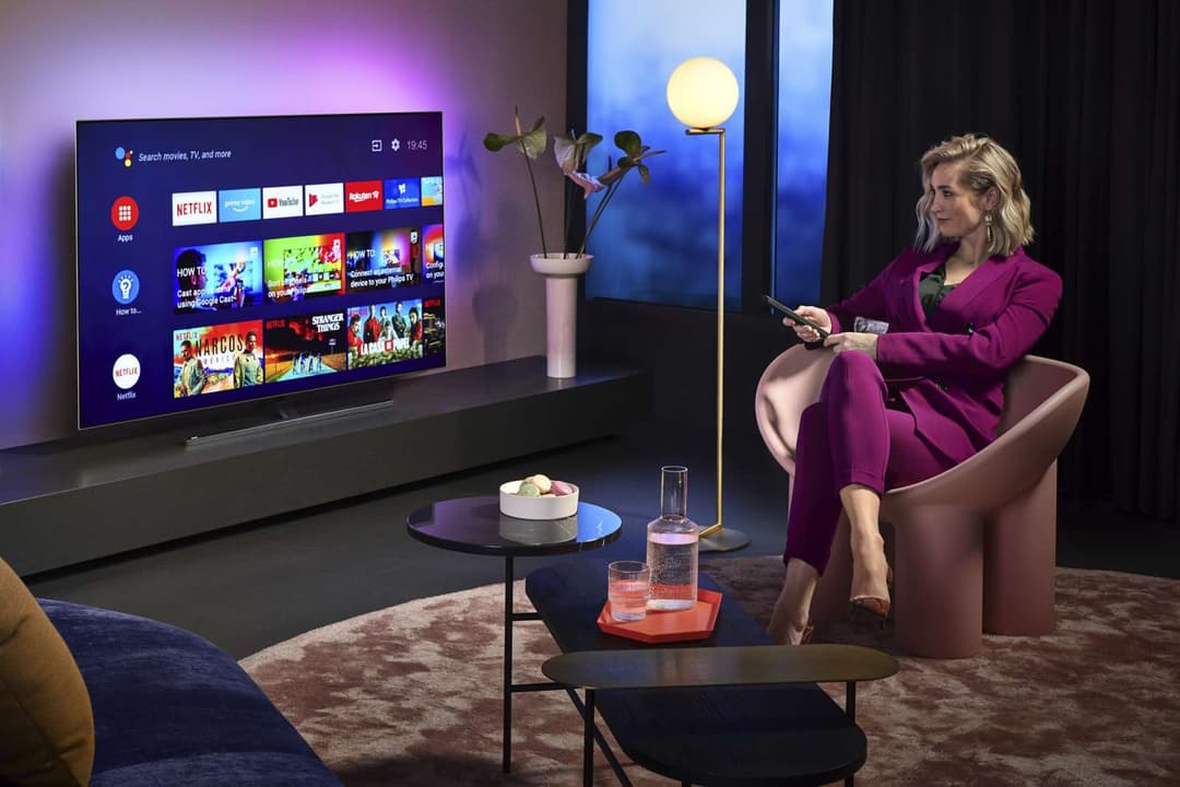 Téléviseur Philips OLED affichant l'interface Android TV dans un salon moderne, femme assise dans un fauteuil rose avec télécommande