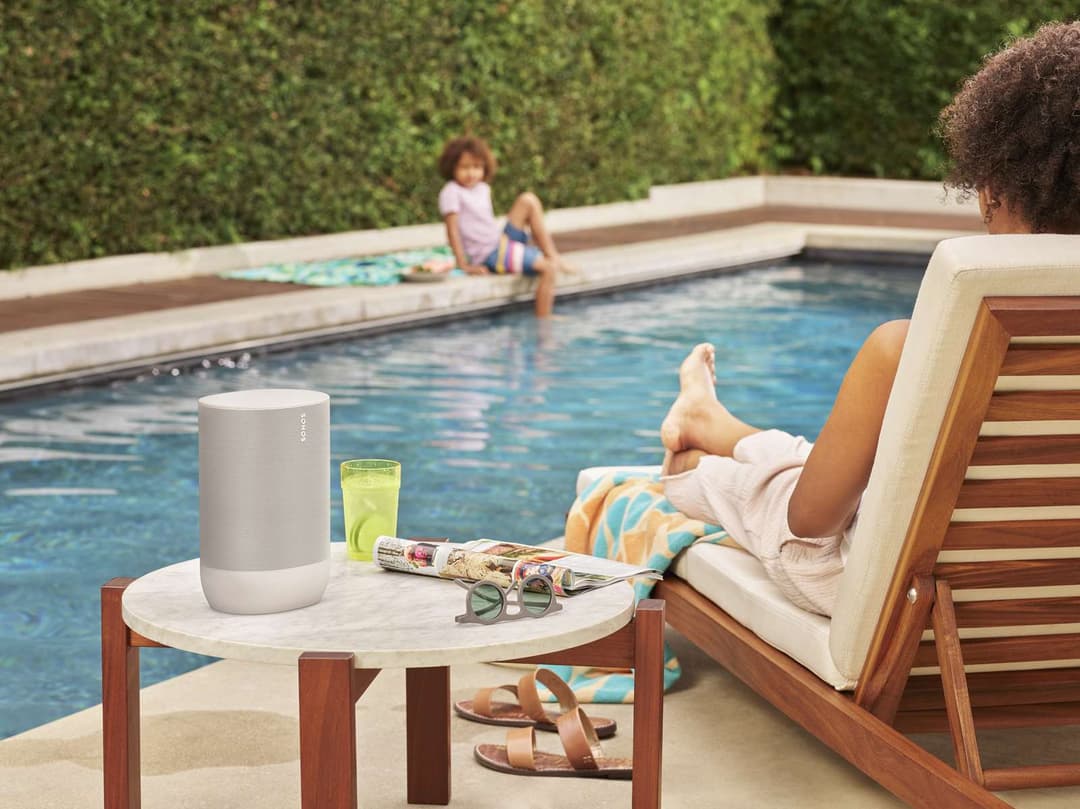 Enceinte Sonos Move blanc crème posée sur une table ronde en bord de piscine, avec chaise longue et paysage aquatique en arrière-plan