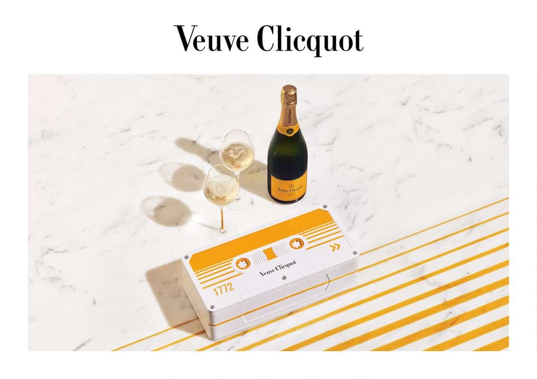 Cassette audio Veuve Clicquot aux teintes blanc et or, posée sur marbre blanc avec champagne et deux flûtes remplies