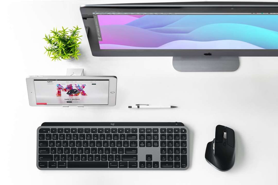 Poste de travail Mac avec écran affichant un design coloré, clavier MX Keys et souris MX Master 3 noirs, petit plant et stylet