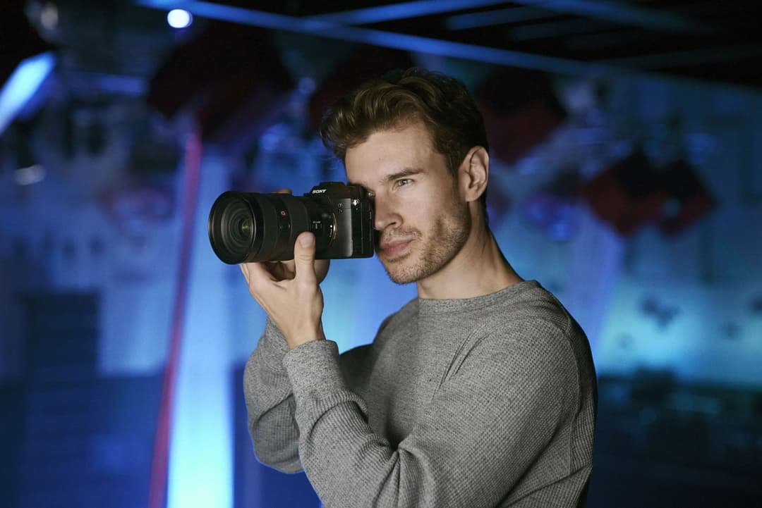 Un homme en pull gris tient un Sony Alpha 7S III face à lui dans un environnement aux lumières bleues et violettes