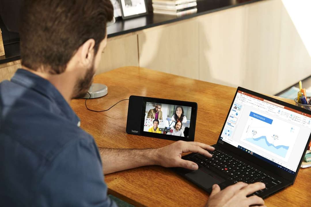 Homme en vidéoconférence sur un Lenovo ThinkSmart View posé sur un bureau en bois, avec un ordinateur portable ouvert