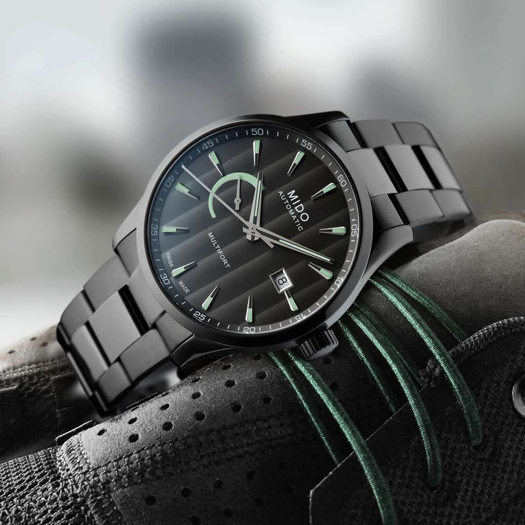 Montre Mido Multifort Power Reserve au cadran noir avec aiguilles et index luminescents, bracelet acier, posée sur un tissu gris