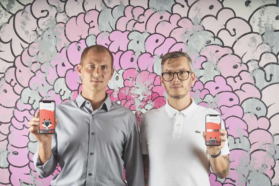 Deux hommes présentent l'application AboutYou sur leurs smartphones devant un mur peint avec des bulles roses et grises stylisées