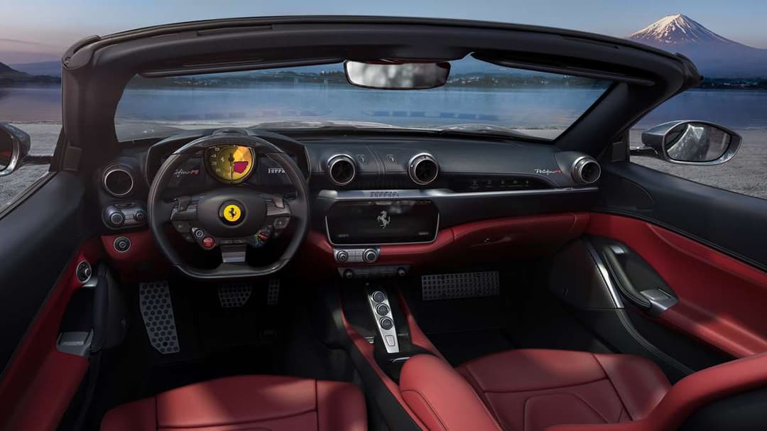 Habitacle de la Ferrari Portofino M décapotable, avec volant et sièges rouges, vue sur un lac et une montagne enneigée