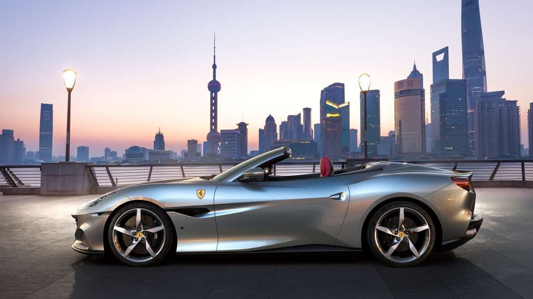 Ferrari Portofino M grise décapotée, vue de profil, stationnée sur une terrasse avec skyline urbaine illuminée en arrière-plan au coucher du soleil