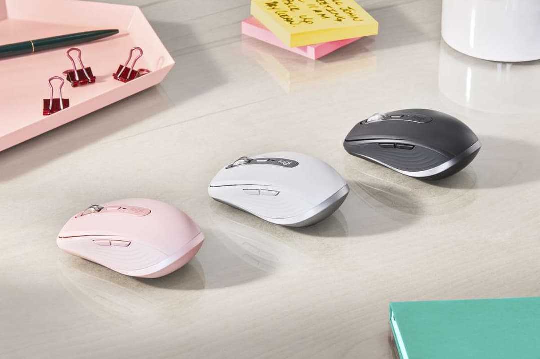 Trois souris Logitech MX Anywhere 3 alignées sur un bureau blanc : rose, blanche et noire, entourées d'accessoires de bureau