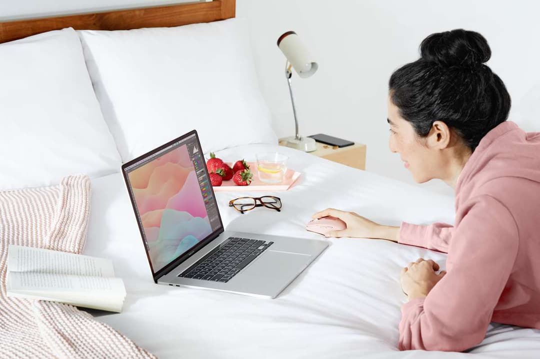 Femme utilisant une souris Logitech MX Anywhere 3 rose sur un lit avec un ordinateur portable