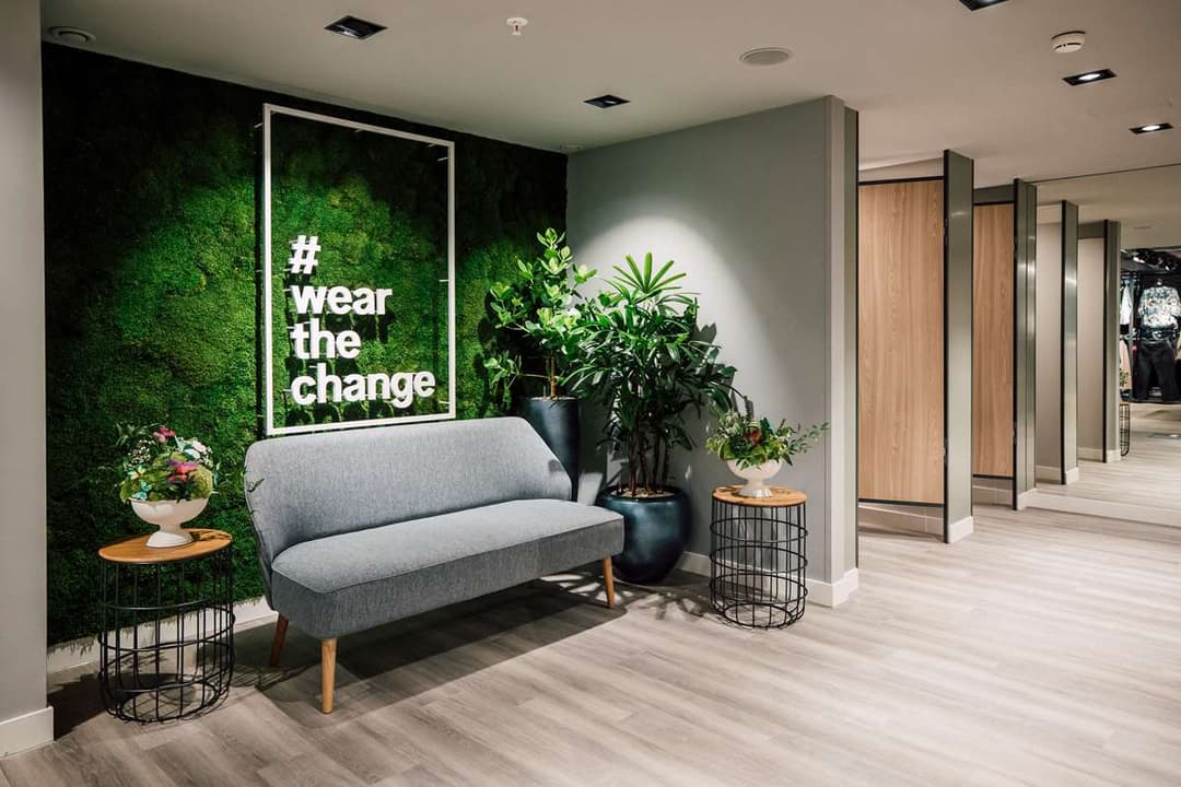 Espace bien-être du magasin C&A avec canapé gris, mur végétal vert, slogan « wear the change », plantes en pots et cabines d'essayage en arrière-plan